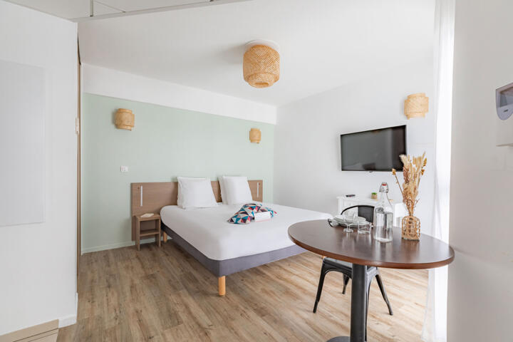 Bright and minimalist Clef Verte certified aparthotel room with a double bed, wicker pendant lamps, a flat-screen TV on the wall, a small dining area with a round table, and natural decorative elements, reflecting an eco-friendly approach to accommodation.