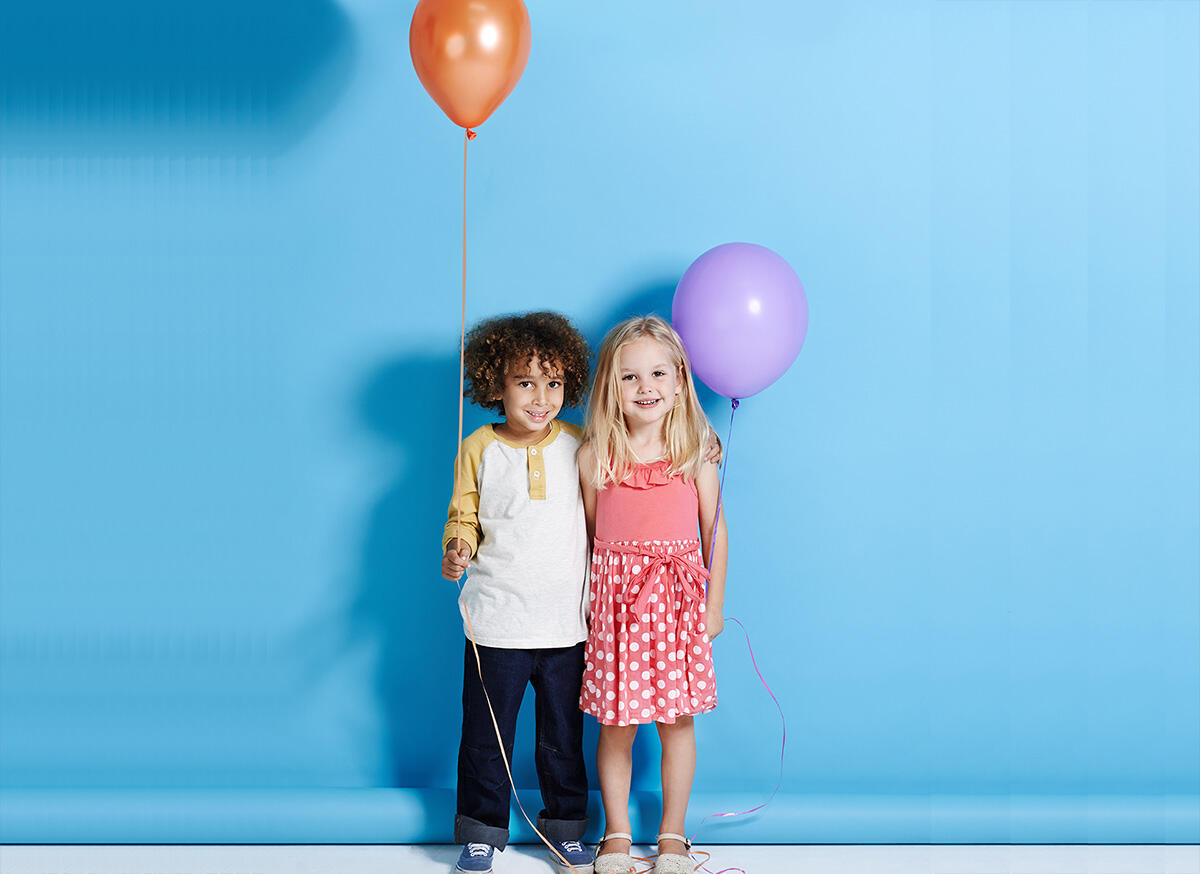Two smiling children holding balloons, a boy with an orange balloon and a girl with a purple balloon, against a blue background for Appart'City's We Love Family offer.