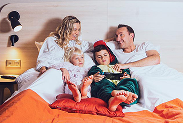 Famille souriante partageant un moment convivial et chaleureux sur un lit, entourée d’une ambiance cosy