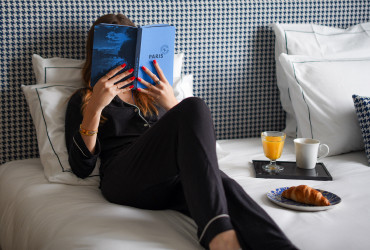 Moment de détente à l’Appart’hôtel Appart’City Paris Roissy CDG, avec une femme lisant un livre sur un lit confortable, accompagnée d’un petit-déjeuner