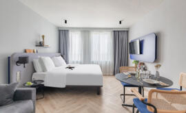 Interior view of an Appart'City Collection Paris Roissy suite with queen-size bed, white bedding, wall-mounted TV, dining area with low table and wicker chairs, and a grey sofa in a bright neutral decor.                            
