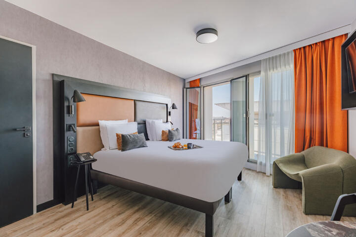 Chambre d'hôtel moderne et confortable avec un grand lit double, des draps blancs, des oreillers décoratifs, un éclairage naturel grâce à une grande fenêtre et des rideaux orange, située à proximité d'une autoroute.