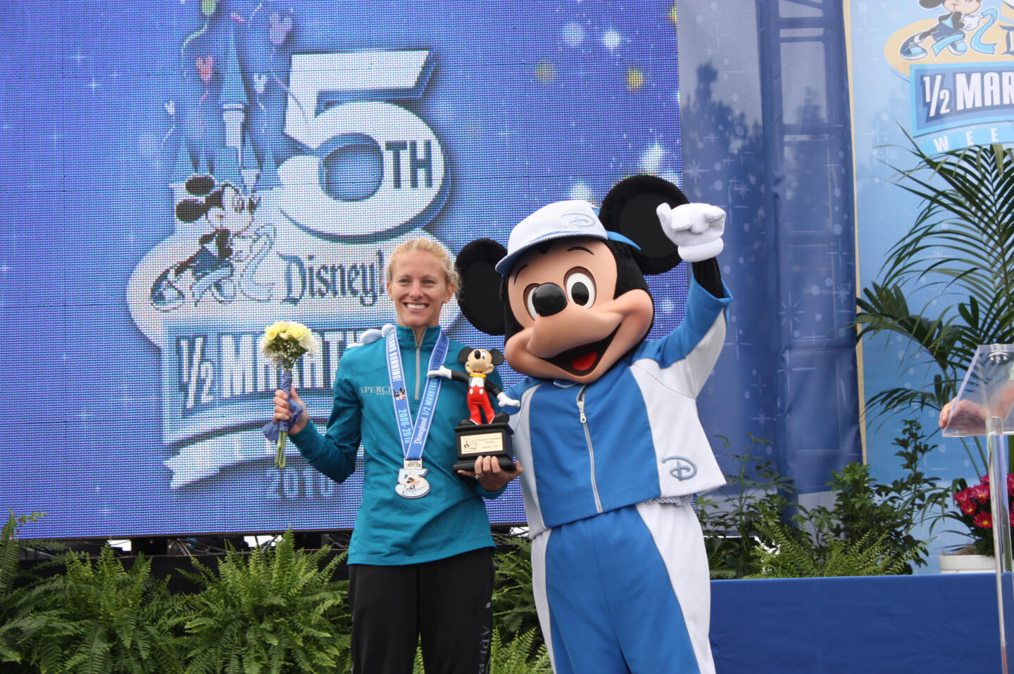Una corredora ganadora sosteniendo un trofeo y un ramo de flores en el escenario, acompañada de Mickey Mouse en ropa deportiva, en la 5ª edición del Disney 1/2 Maratón.