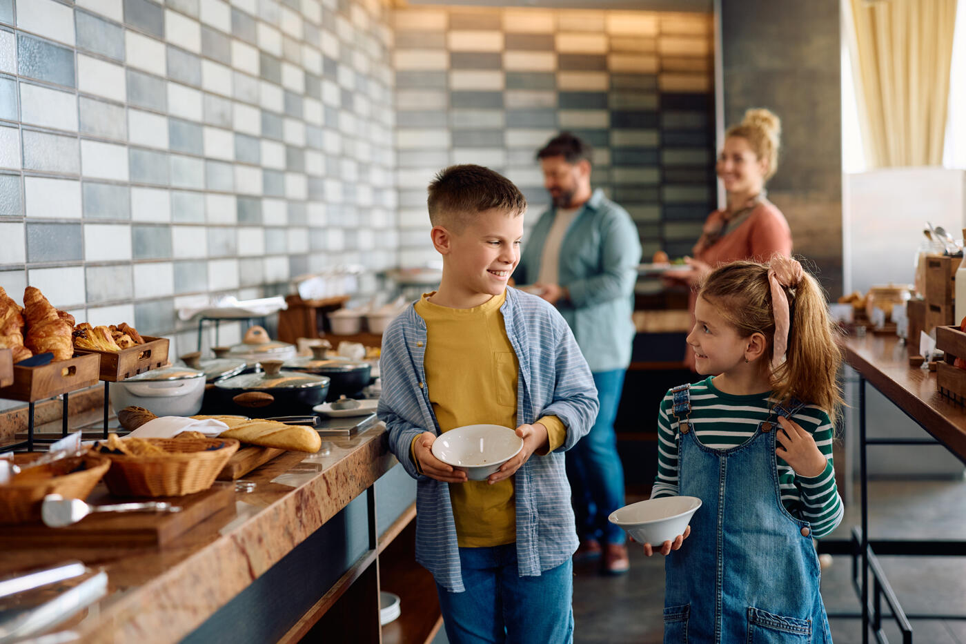 Zwei lächelnde Kinder, ein Junge und ein Mädchen, bedienen sich am Frühstücksbuffet in einem Aparthotel, mit ihren Eltern im Hintergrund.