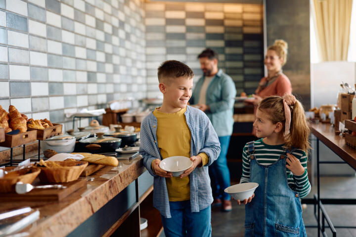 Deux enfants souriants, un garçon et une fille, se servent au buffet du petit déjeuner dans un appart'hôtel, avec leurs parents en arrière-plan.