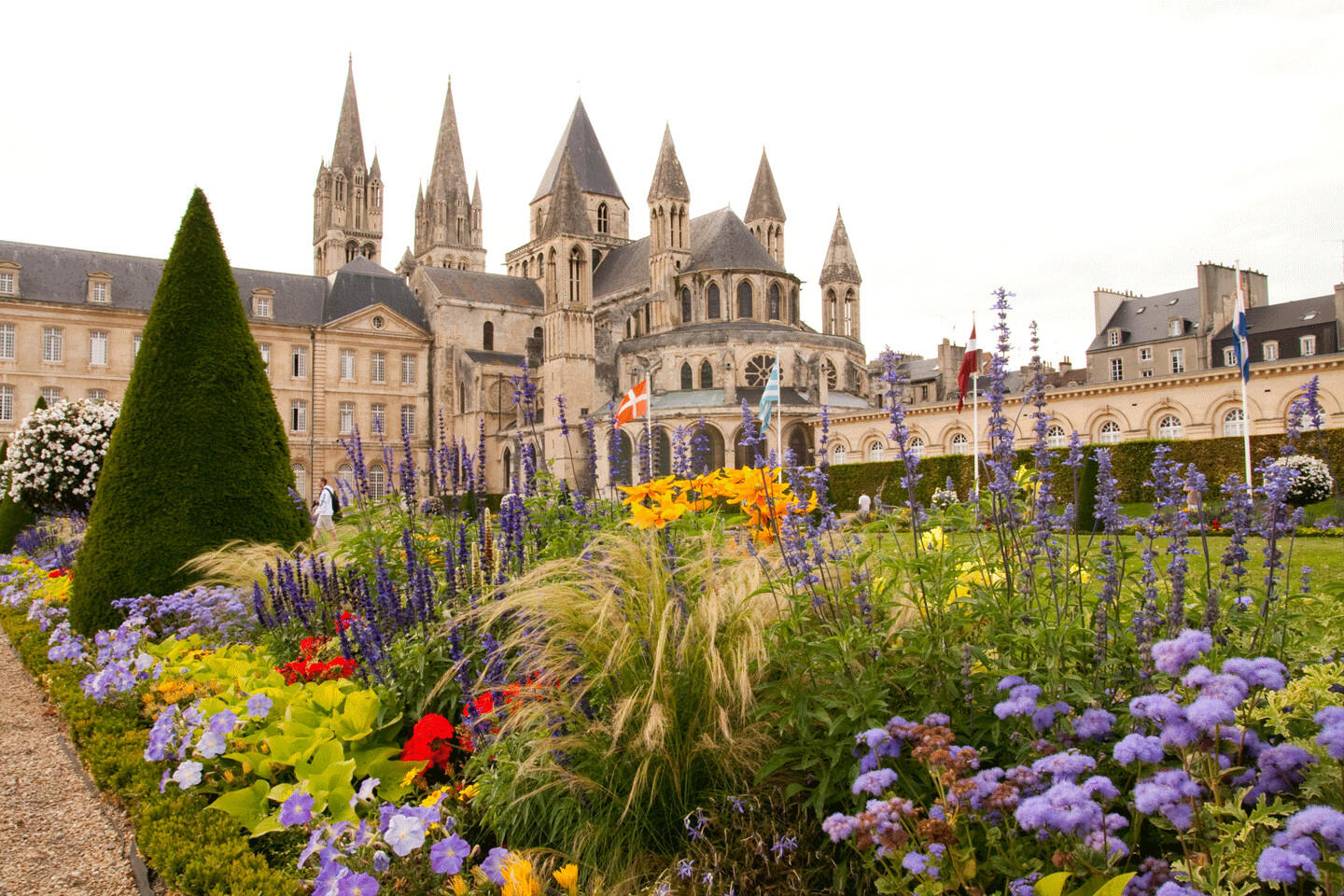 Vue du jardin fleuri et de l'Abbaye aux Hommes à Caen, avec une architecture historique en arrière-plan et un parterre coloré de fleurs en premier plan.