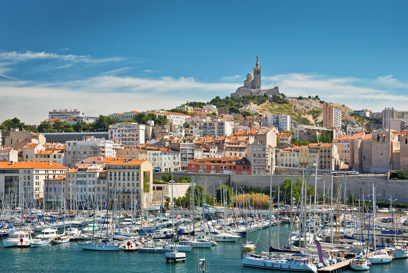 Vue emblématique sur le Vieux-Port de Marseille avec ses voiliers, ses bâtiments historiques et la basilique Notre-Dame de la Garde en arrière-plan.