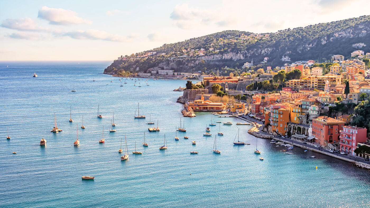 Vue panoramique sur une baie méditerranéenne avec des eaux turquoise, des voiliers ancrés, et une ville côtière colorée entourée de collines verdoyantes.